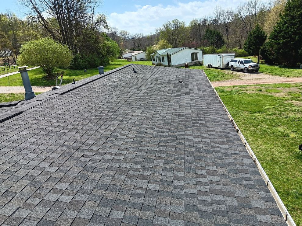 Freshly paved black shingle roof on a house in a rural yard with trees and parked vehicles beyond