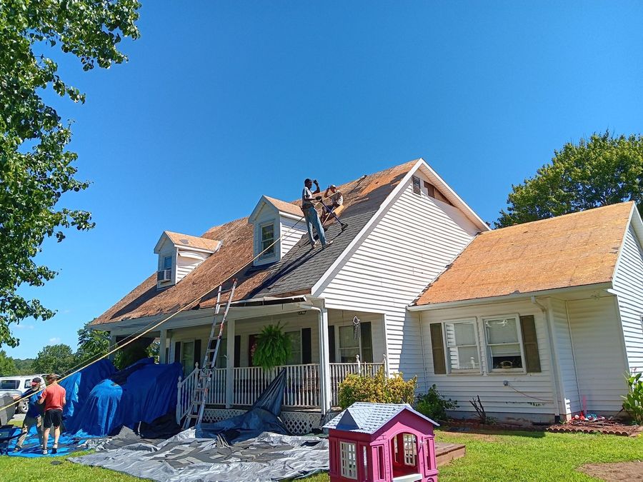 Workers repairing a white house roof on a sunny day, with blue tarp and scaffolding in the yard.