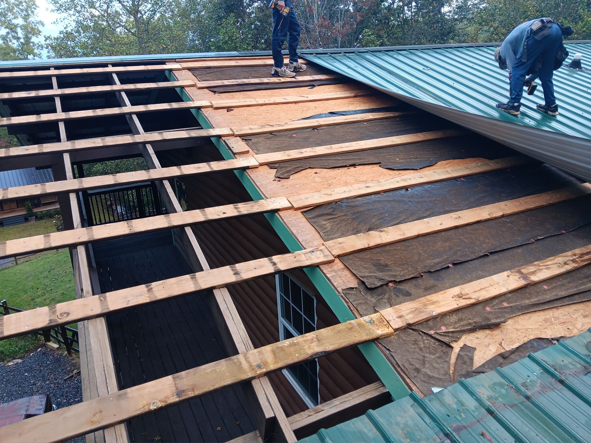 Workers repairing a roof with exposed wooden framing and insulation beneath green metal panels