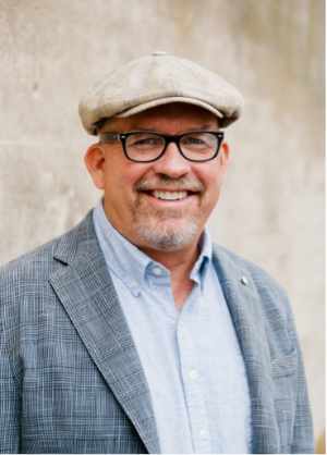 Greg Surrat is wearing glasses and newsboy cap smiling, wearing a blue shirt and gray blazer, standing outside.