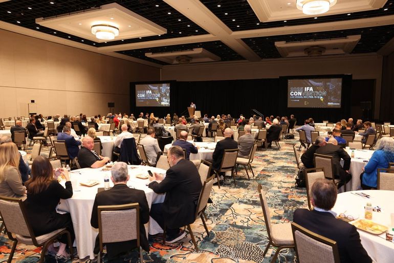 People seated at round tables in a large ballroom, attending a conference, with screens displaying visuals.