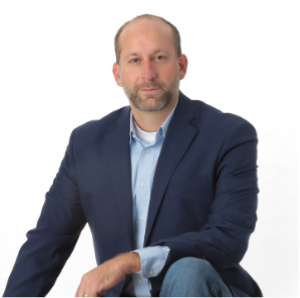 Joe Soltis in blue blazer, shirt and jeans, smiles while kneeling.
