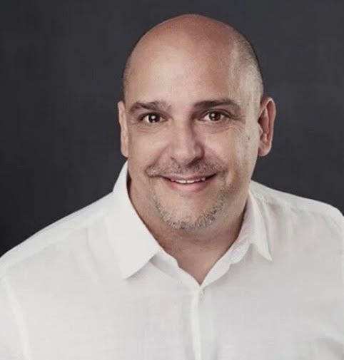 Joe Mohay with a slight smile wearing a white button-down shirt. Dark gray background.