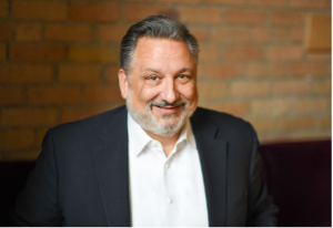 John Francis with graying hair and beard in a suit jacket, smiling, against a brick background.