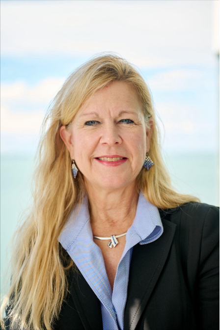 Kristi Mailloux is smiling, wearing a black blazer, light blue shirt, and jewelry; sea and sky background.