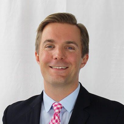 Michael Layman is wearing a suit smiling at the camera, wearing a pink checkered tie. White background.