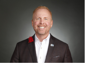 Red Boswell is smiling, wearing a suit jacket and white shirt. Red flower on lapel.