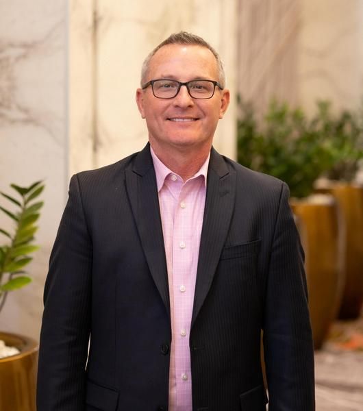 Sean Falk in glasses, dark suit, pink shirt smiling at the camera indoors.