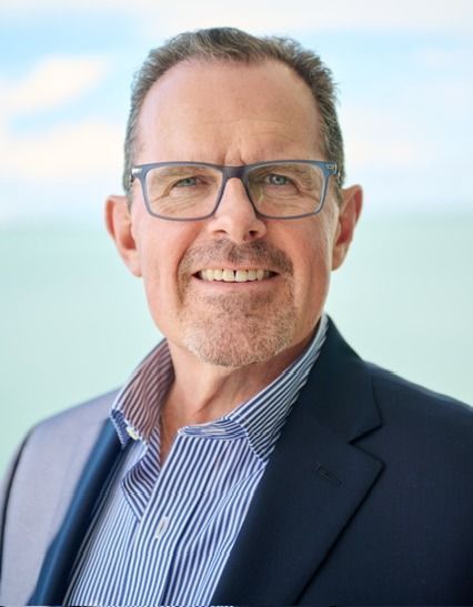 Todd Recknagel with glasses, smiling, wearing a blue striped shirt, and navy blazer in a sunny outdoor setting.
