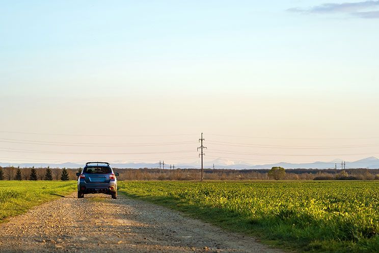 Blue car on a gravel road beside green fields under a pale sky