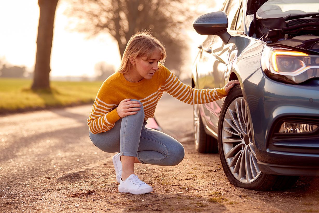 Woman squats beside a blue car with the hood open, examining the front tire on a rural road.