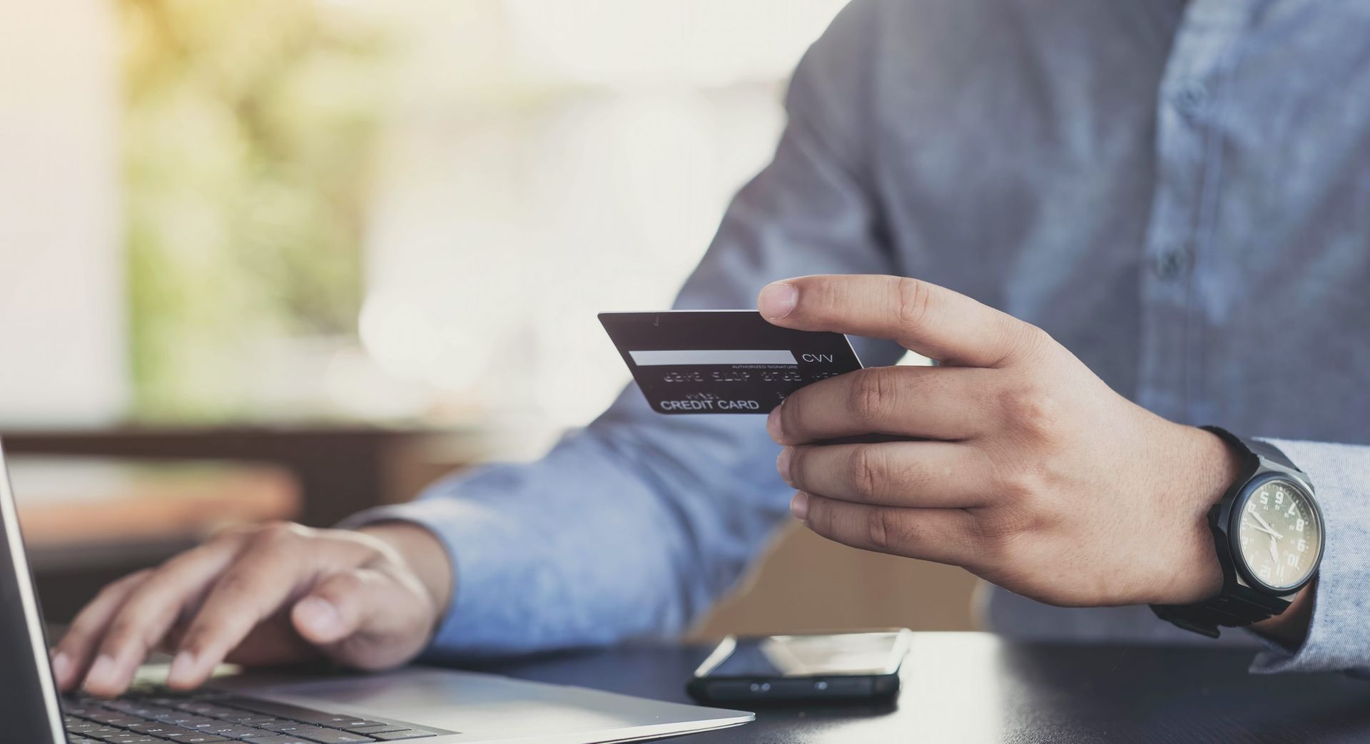 A person in a blue shirt holds a credit card while using a laptop to make an online purchase.