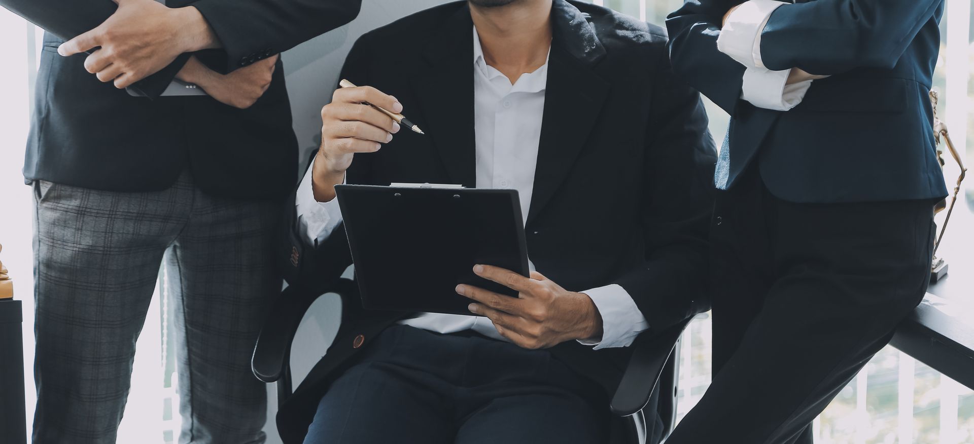 People in business suits in an office, one seated and holding a pen and clipboard, others standing.