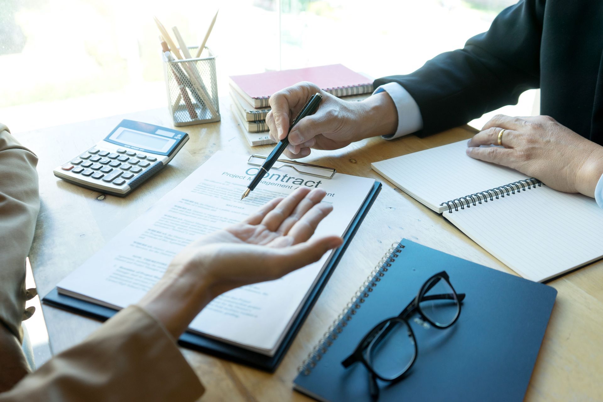 Two people sit at a table reviewing a contract with a pen, calculator, notebooks, and glasses nearby.