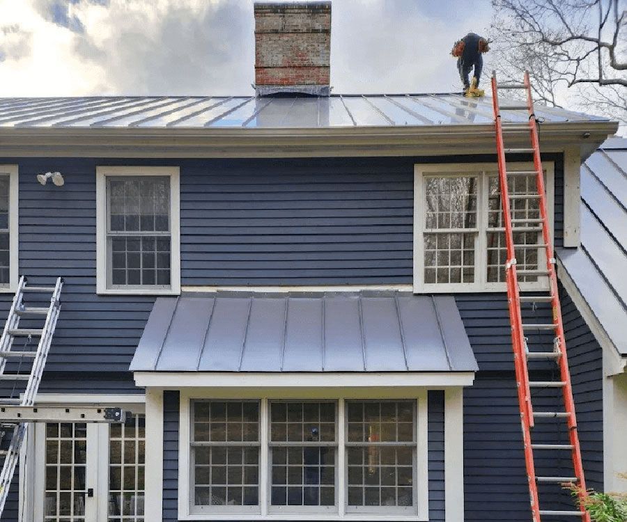 A worker stands on a dark-blue house's metal roof near a brick chimney, with ladders positioned against the exterior.