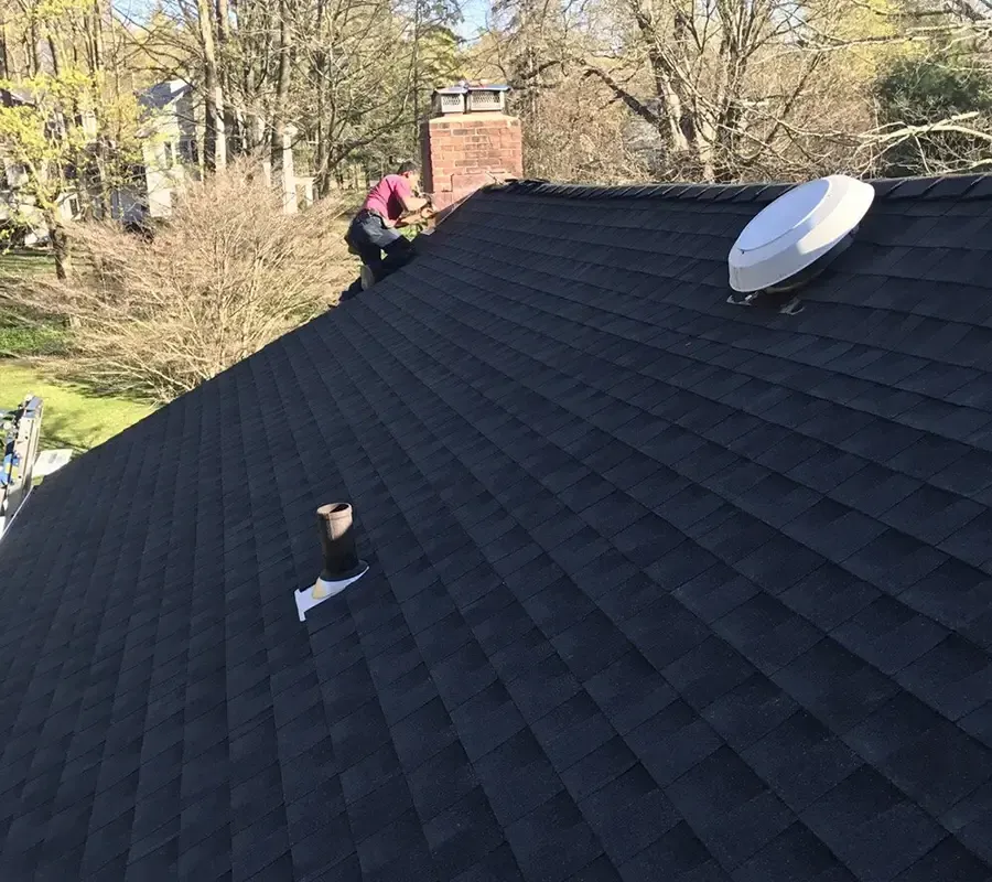 A person in red working on a dark shingled roof near a chimney, with a white satellite dish installed nearby.