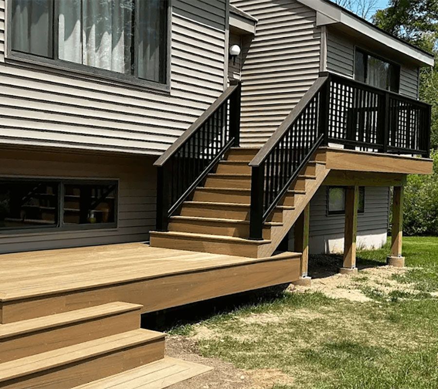 A tan composite deck with a set of stairs leading to an upper level, featuring black metal railings against gray siding.