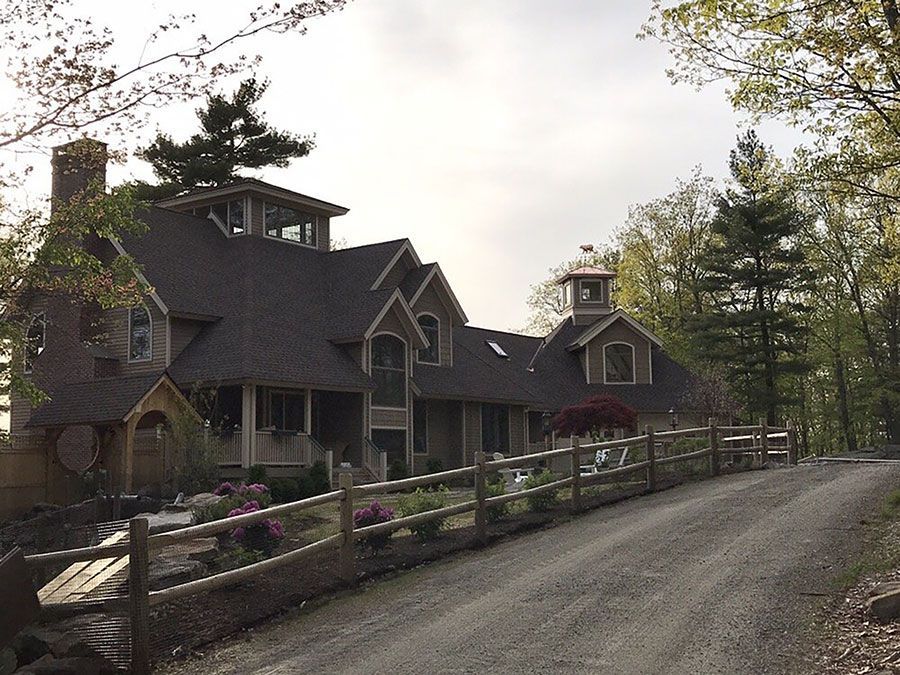 A multi-level home with dark roofing and natural siding, viewed from a gravel driveway behind a wooden fence.