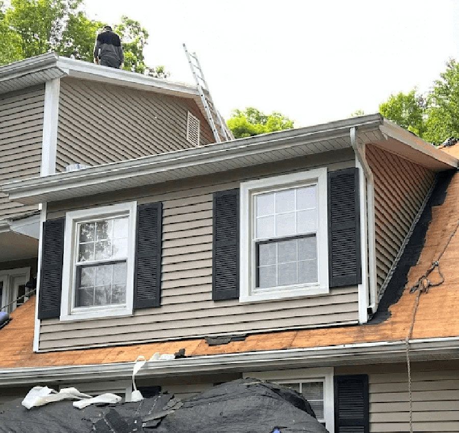 A person working on a residential roof during a renovation, showing a two-story house with exposed wood sheathing.