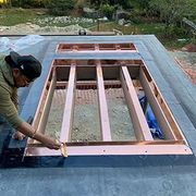 A worker installs copper flashing around a rectangular roof opening for a skylight on a flat, dark-surfaced roof.