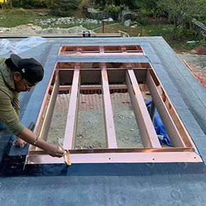 A worker installs copper flashing around a rectangular roof opening for a skylight on a flat, dark-surfaced roof.