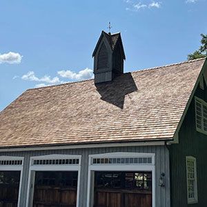 A green and grey wooden garage with a shingled roof and a decorative wooden cupola under a blue sky.