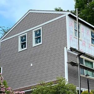 Side view of a house undergoing renovations, showing grey siding, windows, and exposed house wrap on the upper section.