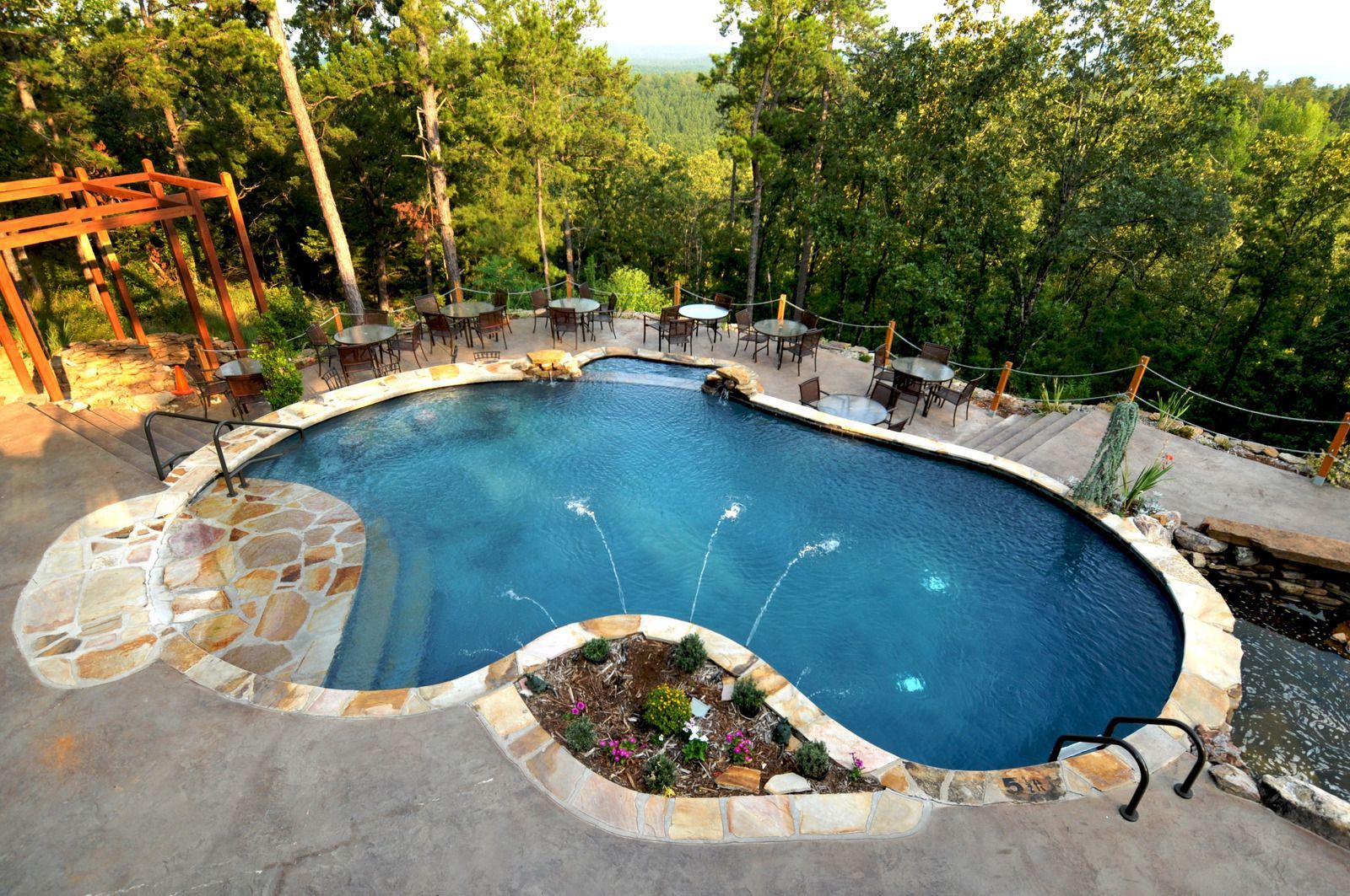 Pool with waterfall, fountains, surrounded by stone patio and tables, in a forest setting.
