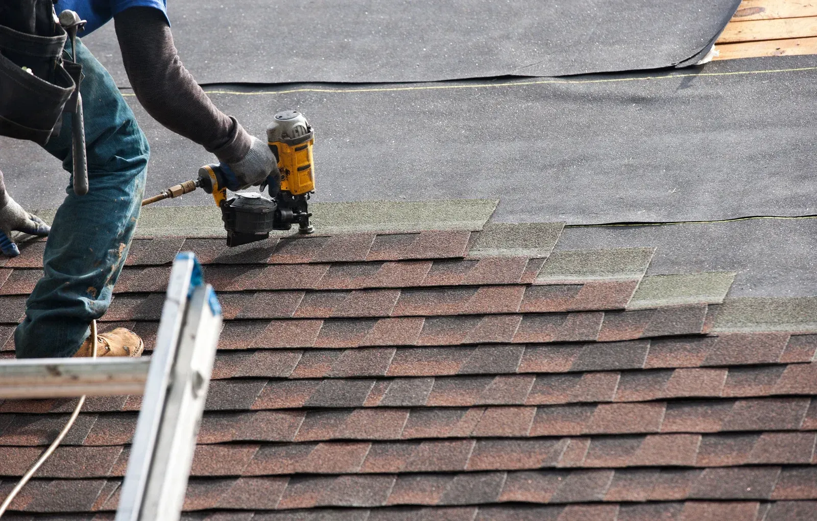 Roofer using a nail gun to install brown asphalt shingles on a roof.