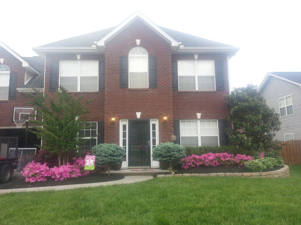 Two-story brick house with pink flowers, green grass, and black shutters; daytime.