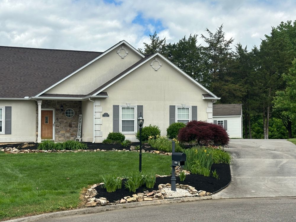 Beige house with gray shutters, brown roof, and landscaped yard on a cloudy day.