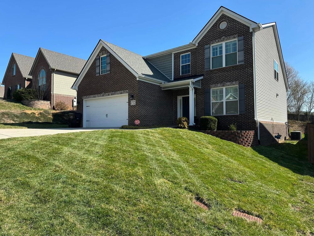 Two-story brick house on a slight hill with green lawn and blue sky. Another house visible in the distance.