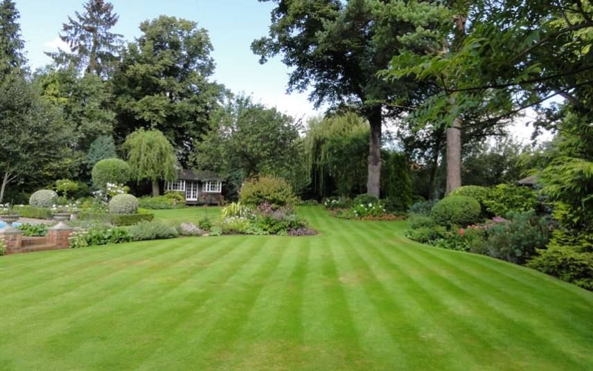 Lush green lawn with striped mowing pattern, garden beds, and trees. Small building in the distance.
