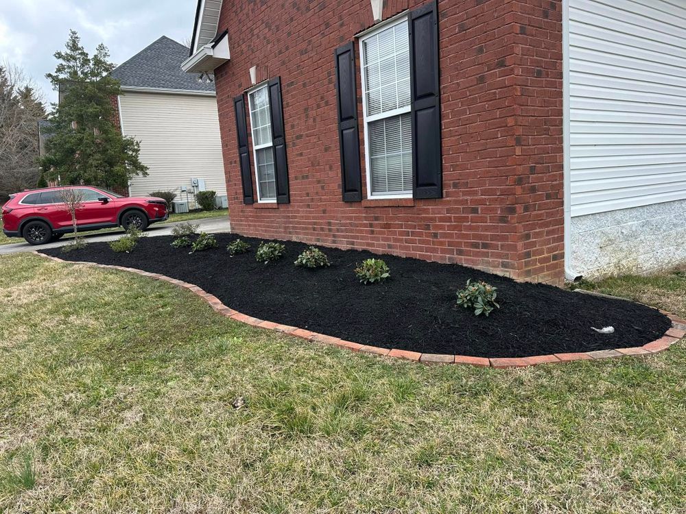 A brick house with black shutters and a red car parked in the yard. Mulched flower bed.