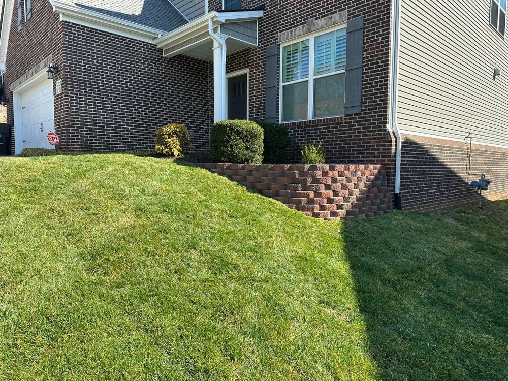 Brick house with a retaining wall on a grassy hill. Bushes and a window are visible.