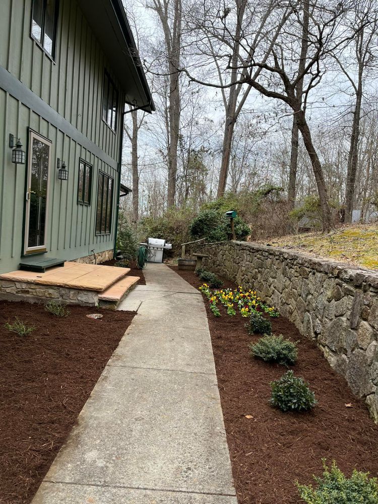 Side of a green house with a concrete pathway, lined with landscaping and a stone wall.