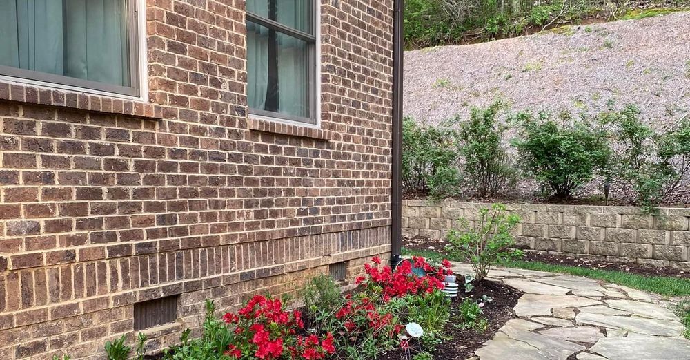 Brick house exterior with red flowers, stone path, and a retaining wall on a hillside.