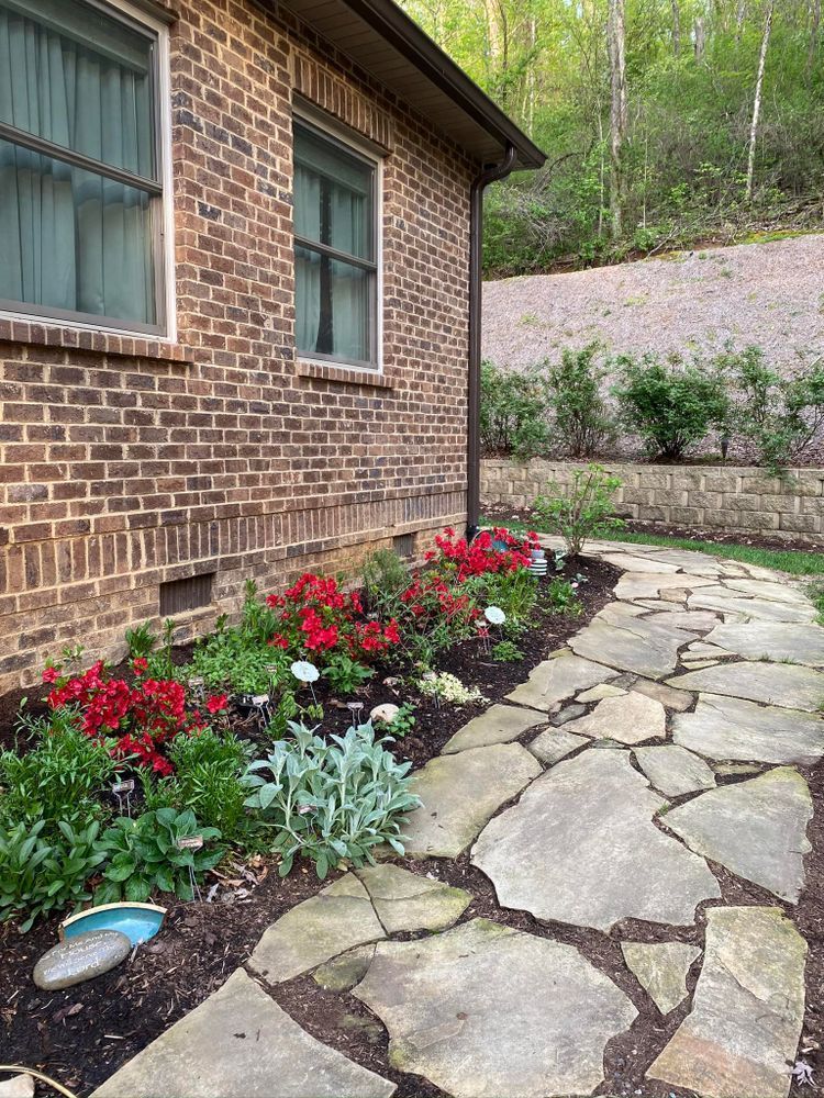 Brick house with flower bed, stone path; red blooms, green plants, hillside in background.