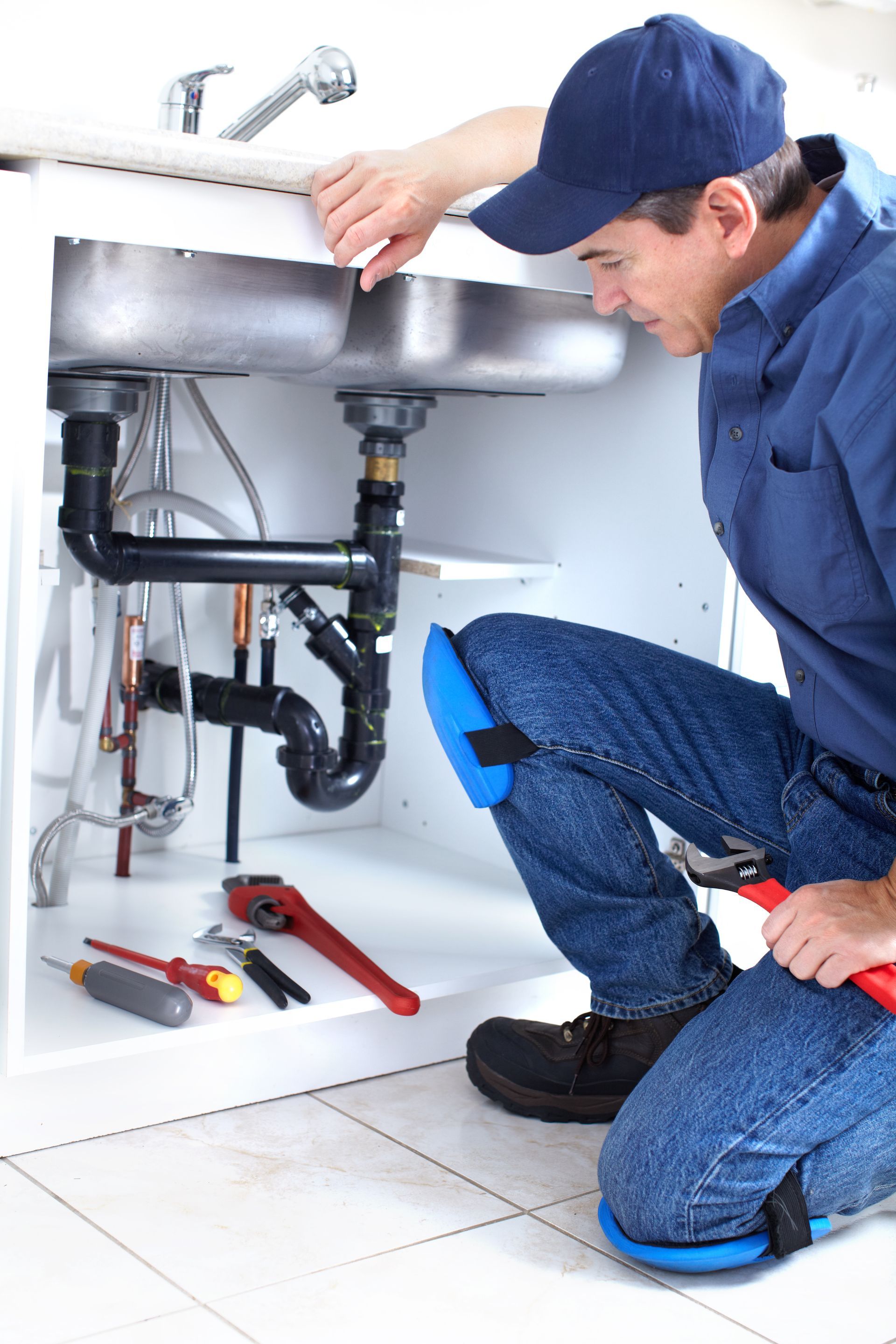 A worker for local plumbing contractors kneels to repair complex piping under a double kitchen sink.