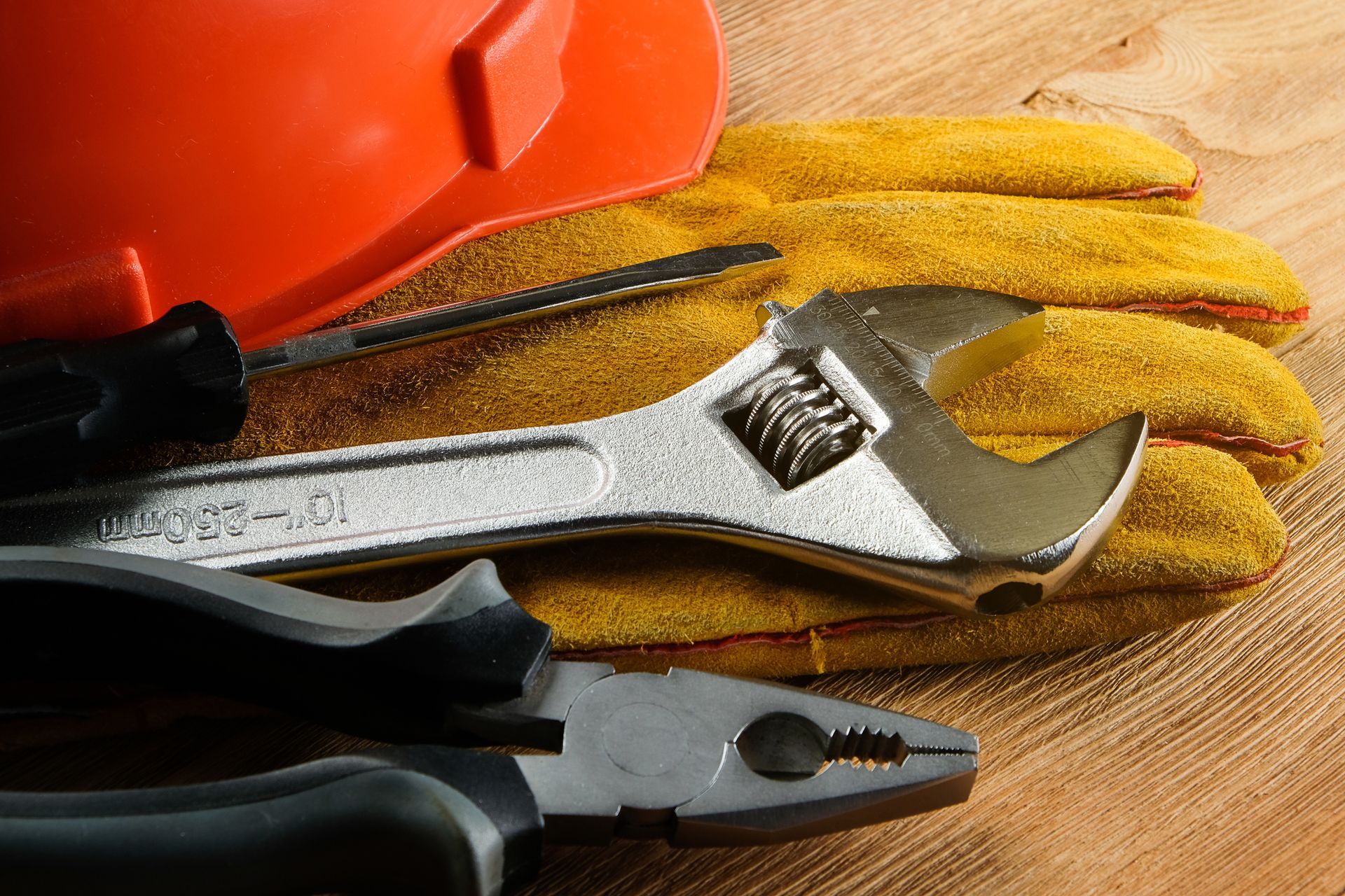 Orange hard hat, work gloves, wrench, screwdriver, and pliers on a wooden surface.