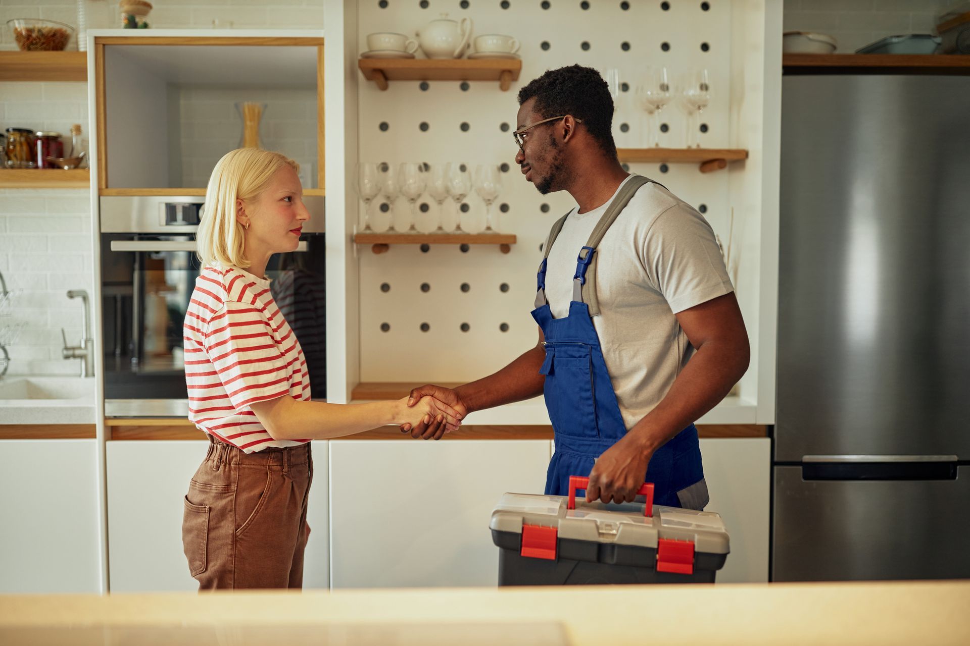 Woman shaking hands with a repair person in a kitchen. The repair person holds a toolbox.