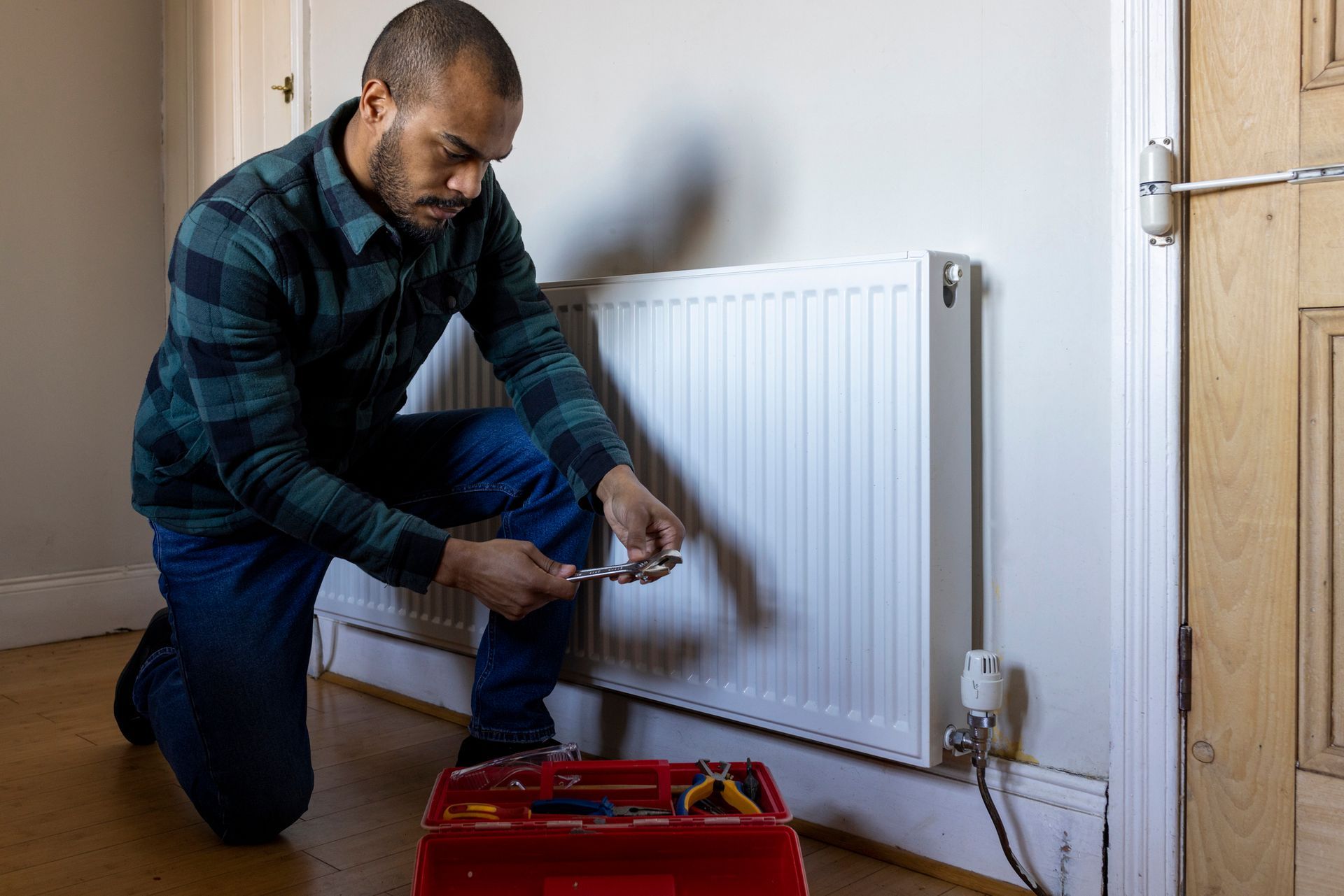 Man kneels to work on radiator, with tools in a red toolbox. He wears plaid shirt and jeans, indoors.