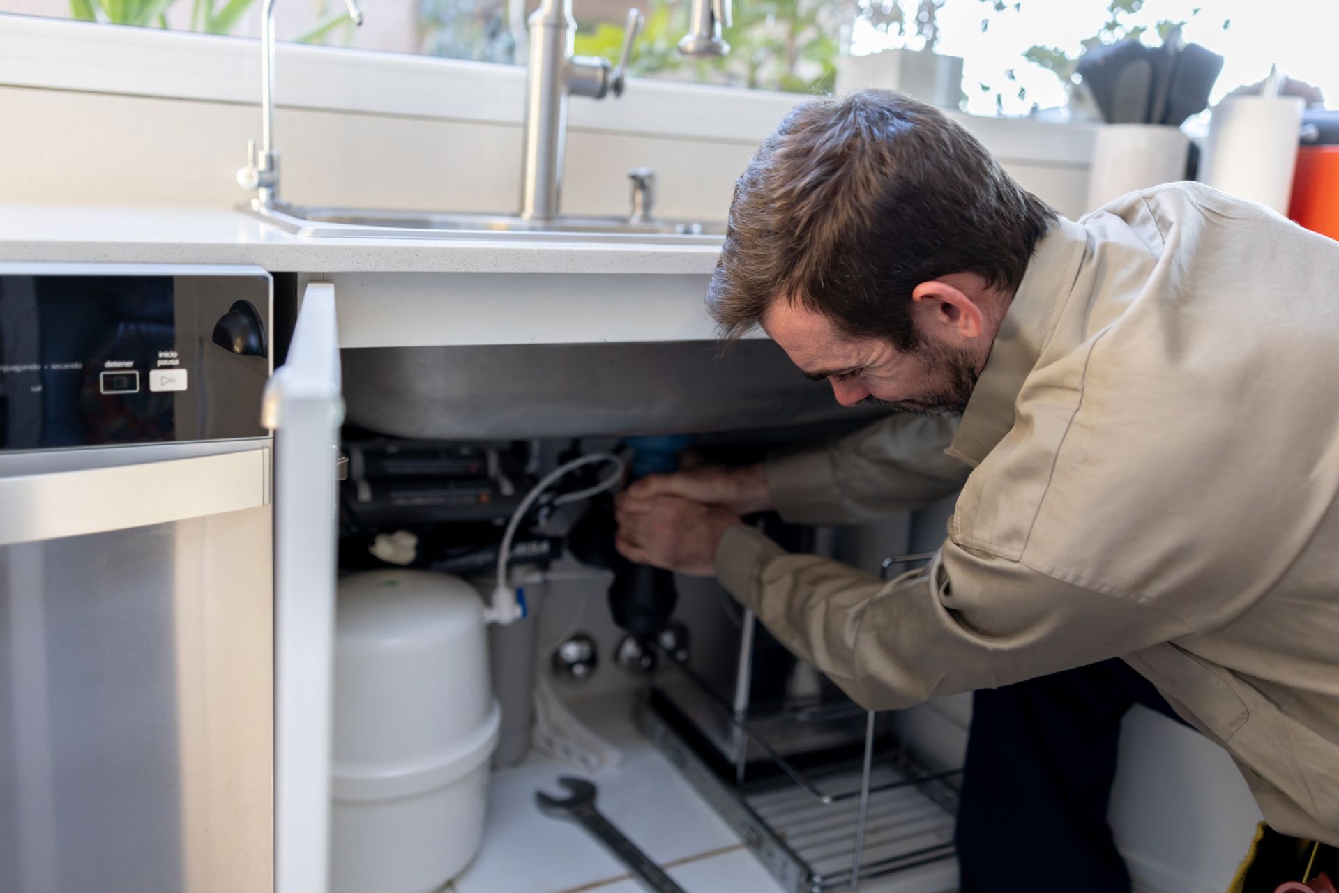 Plumber repairs pipes under a kitchen sink. He wears tan work shirt, uses a wrench, and appears focused.
