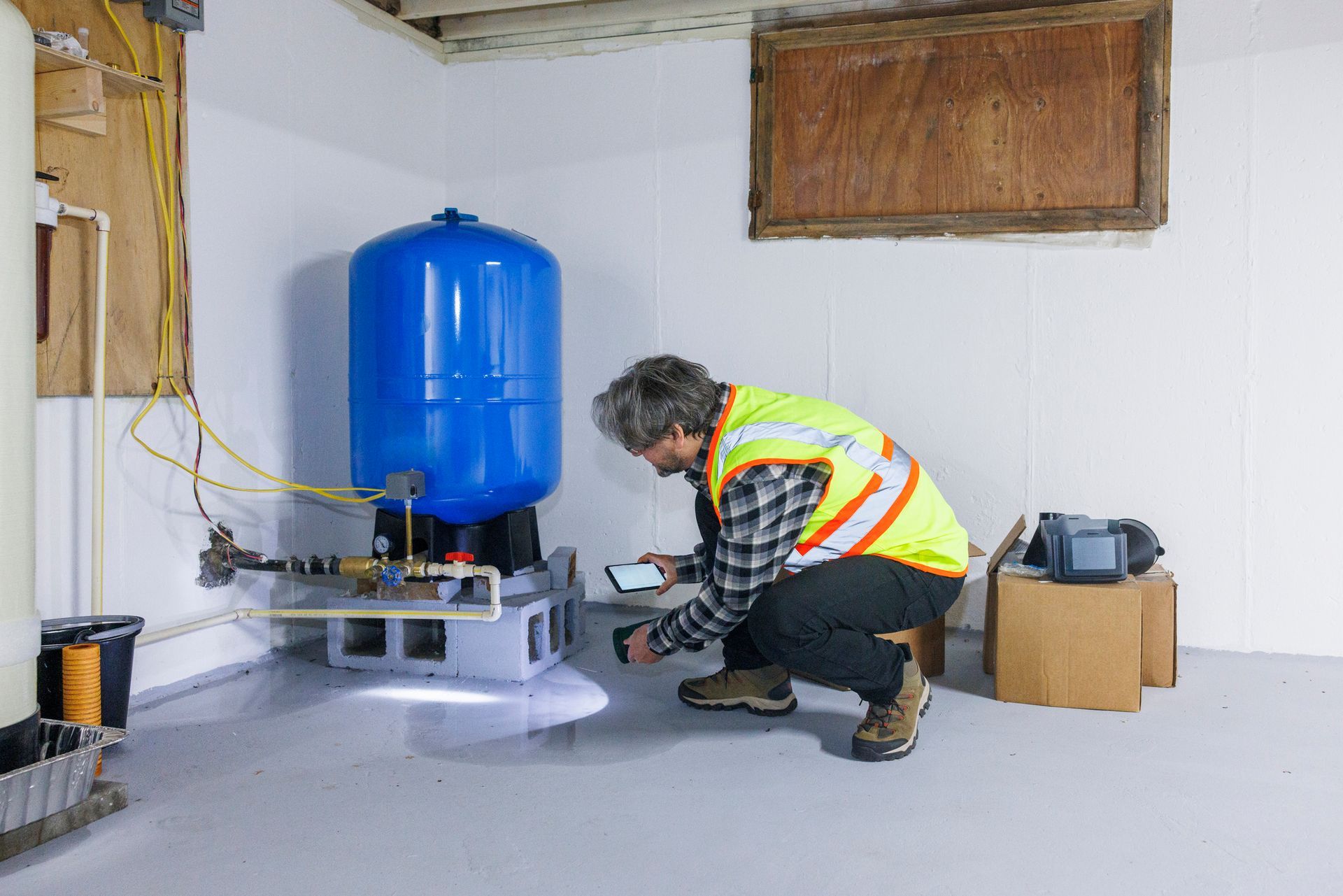 Man in safety vest examines a water pump in a basement, using a phone. Blue tank, white walls, and gray floor.