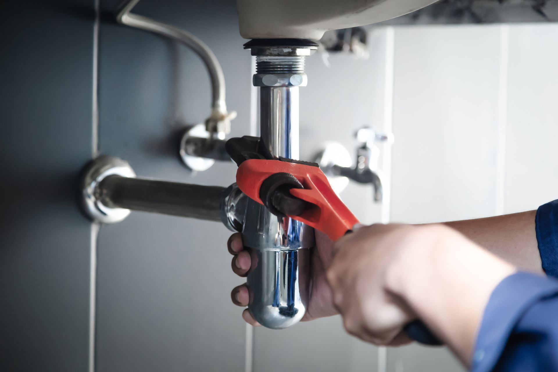 Plumber using an orange wrench to work on pipes under a sink.