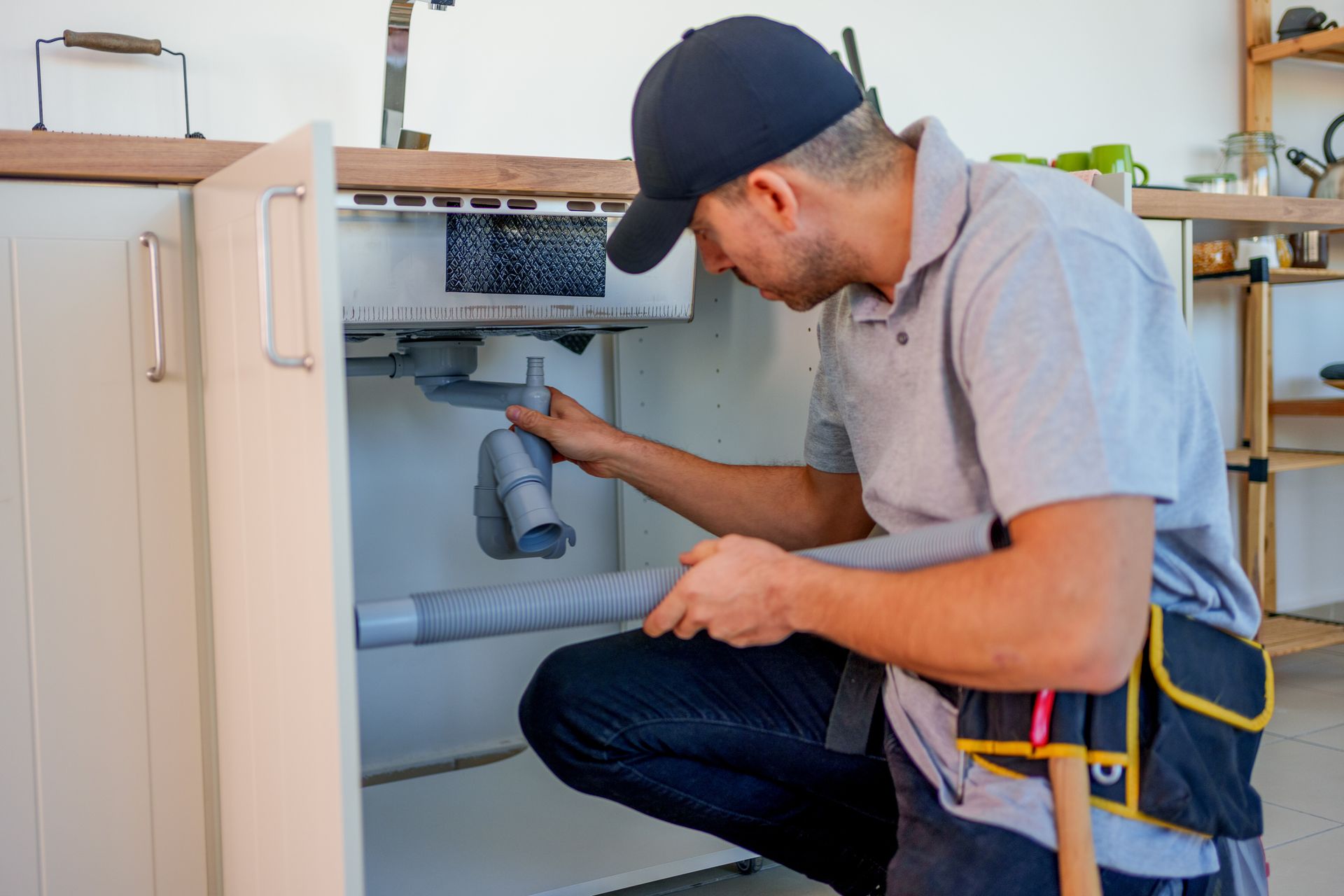 Plumber examines pipes under a kitchen sink. He wears a cap, polo shirt, and tool belt.