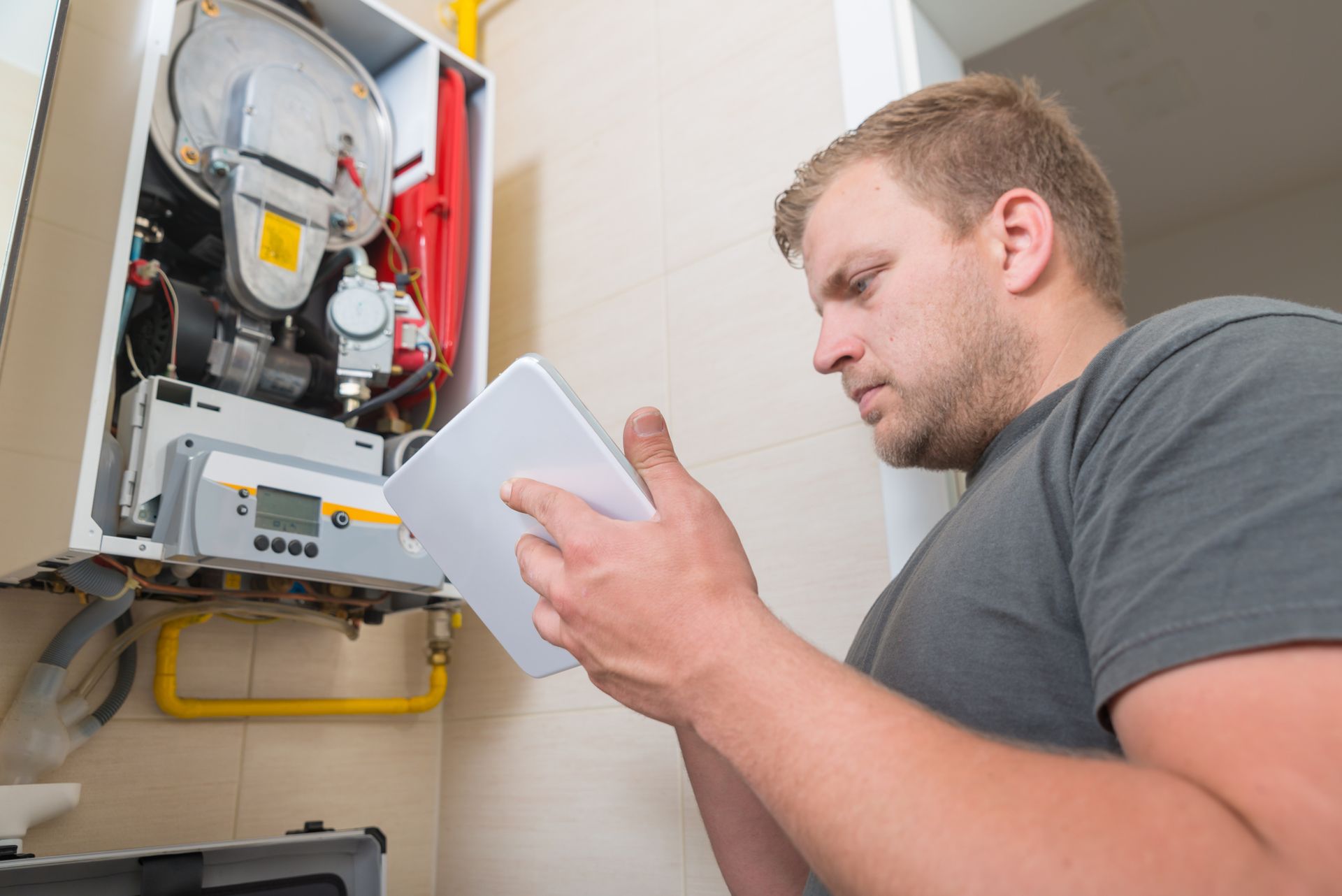 Man examining a heating system control panel, holding a tablet, inside a home.