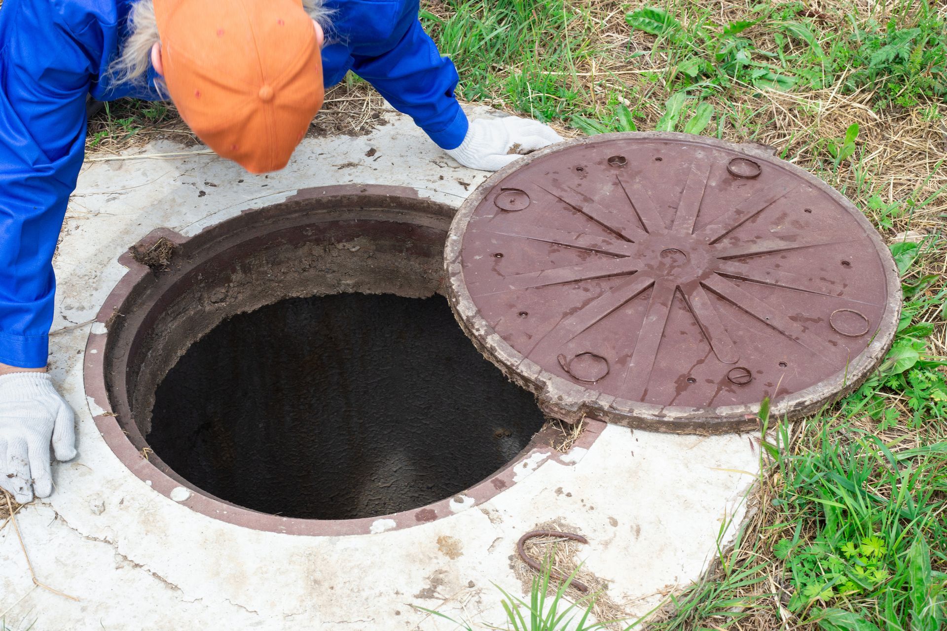 Person in blue coveralls and orange hat, looking into an open manhole. The metal lid is partially removed, and the ground is grassy.