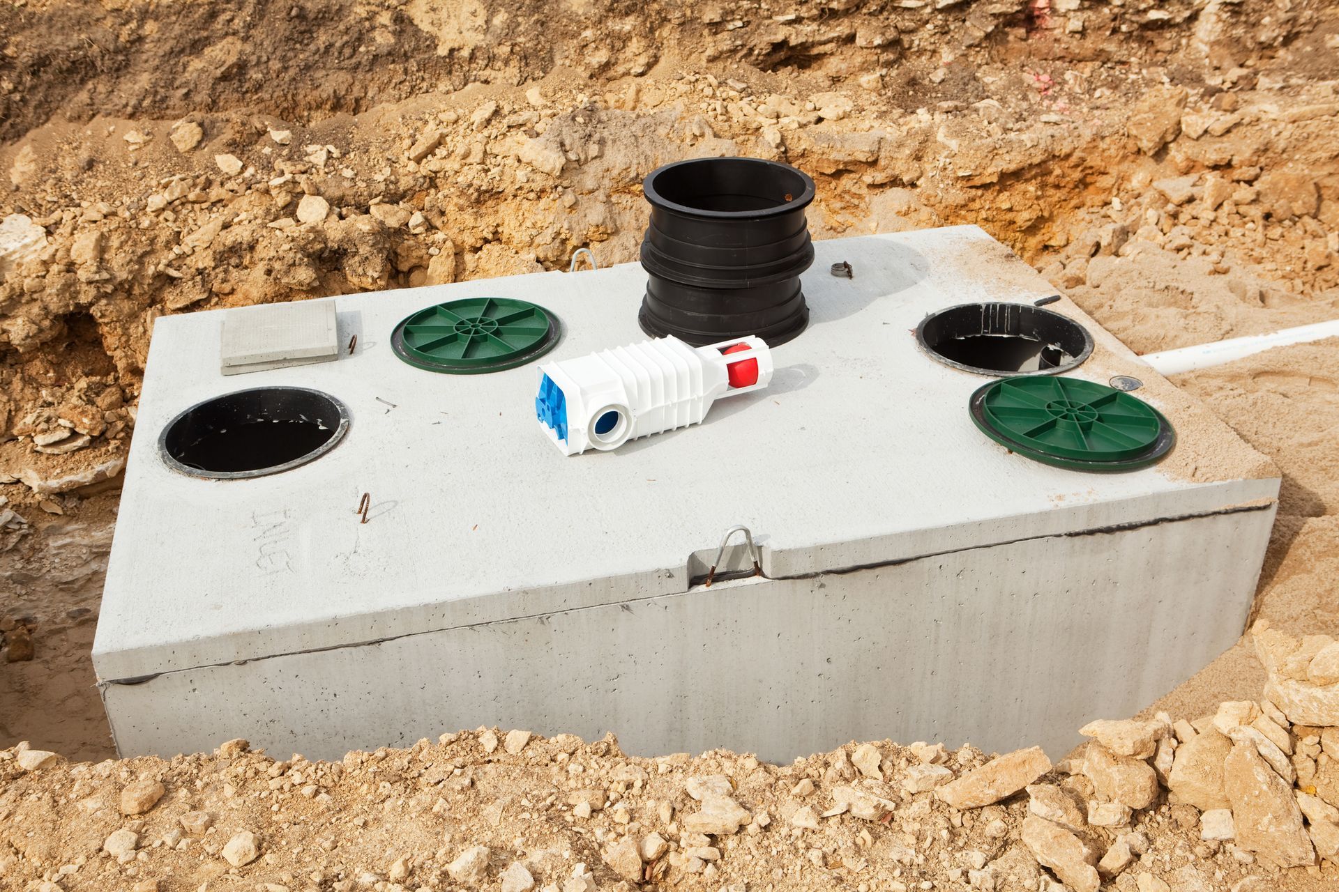 A concrete septic tank, partially buried, with access lids, pipes, and a white and red fitting.