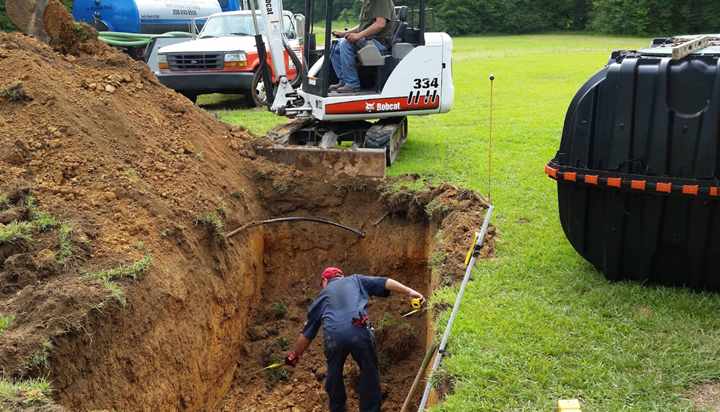 A worker in a deep trench, using a shovel. A small excavator and septic tank are nearby in a grassy area.